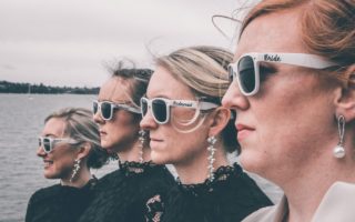 Stylish bride and bridesmaids in sunglasses standing on sea embankment on wedding day