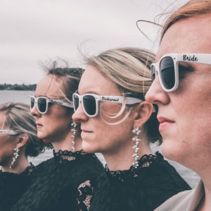 Stylish bride and bridesmaids in sunglasses standing on sea embankment on wedding day