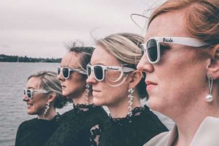 Stylish bride and bridesmaids in sunglasses standing on sea embankment on wedding day