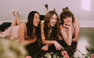 Three girls are wearing AW Bridal bridesmaid dresses and laughing happily in the room. There are also some flowers on the ground.