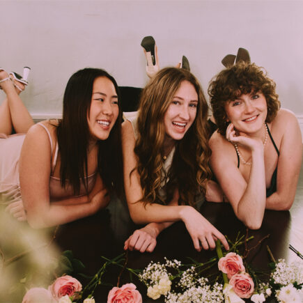 Three girls are wearing AW Bridal bridesmaid dresses and laughing happily in the room. There are also some flowers on the ground.