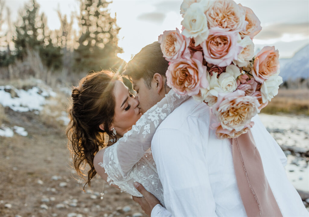 The bride holds a bouquet in her hands and kisses her lover passionately in the sun.