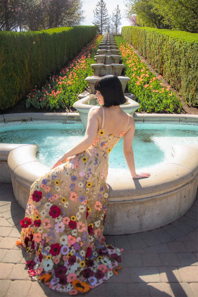 A short-haired woman in a floral maxi dress lounges gracefully by a fountain, with a beautiful tulip bed in the background, her eyes closed in enjoyment.