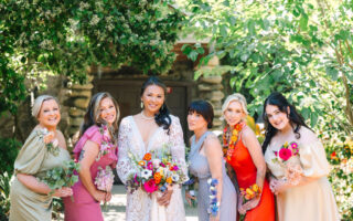 A group of beautiful women, holding flowers, are smiling radiantly in the sunlight.