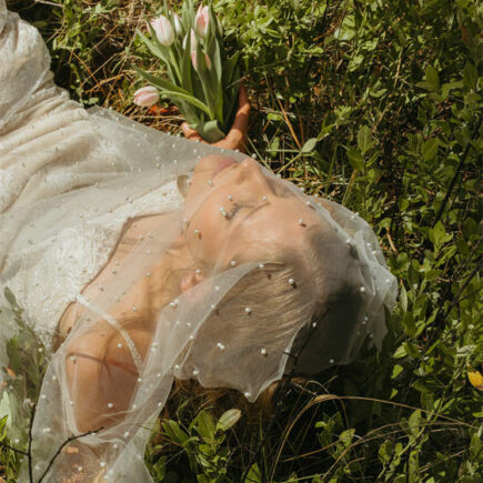 Woman in a wedding dress lying in grass, holding a bouquet of pink tulips.