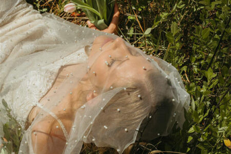 Woman in a wedding dress lying in grass, holding a bouquet of pink tulips.