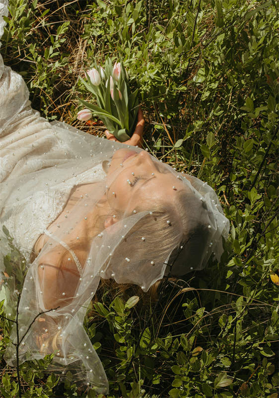 Woman in a wedding dress lying in grass, holding a bouquet of pink tulips.