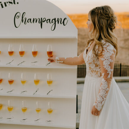 A bride standing next to rows of champagne flutes, holding one in her hand.