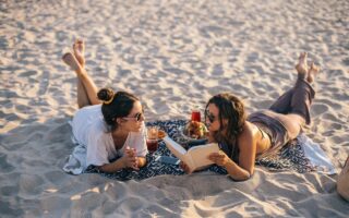 Two women lying on a blanket at the beach, one reading a book and the other listening.