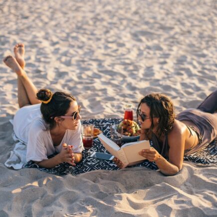 Two women lying on a blanket at the beach, one reading a book and the other listening.