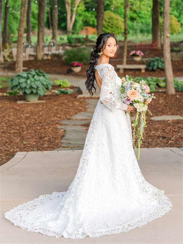 A bride in a lace wedding gown holding a bouquet, standing on a pathway in a garden with trees and plants in the background.