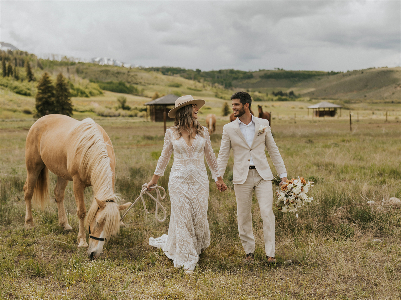 A bride in a Bohemian-style wedding gown and hat holds hands with the groom while leading a horse in a grassy field.