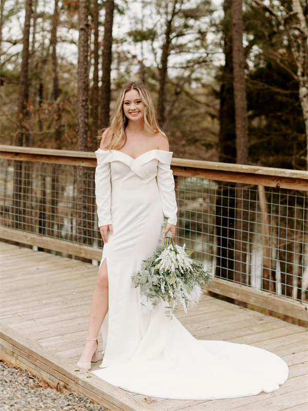 A bride in a satin off-shoulder wedding gown stands on a wooden bridge holding a bouquet of greenery.