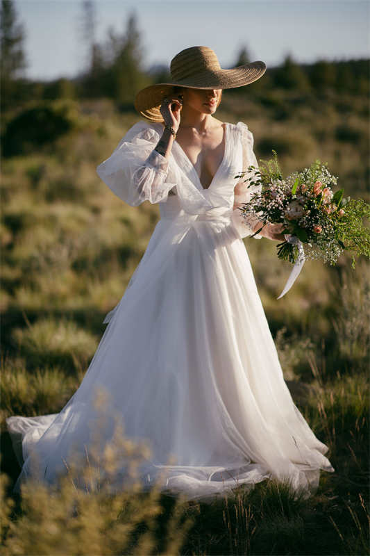 A bride in a tulle wedding gown with puffed sleeves and a wide-brimmed hat, holding a bouquet in a field.