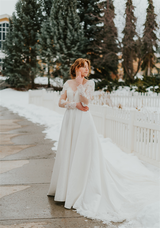 A woman in a white wedding dress with lace sleeves standing on a snowy pathway next to a white fence, with evergreen trees in the background.