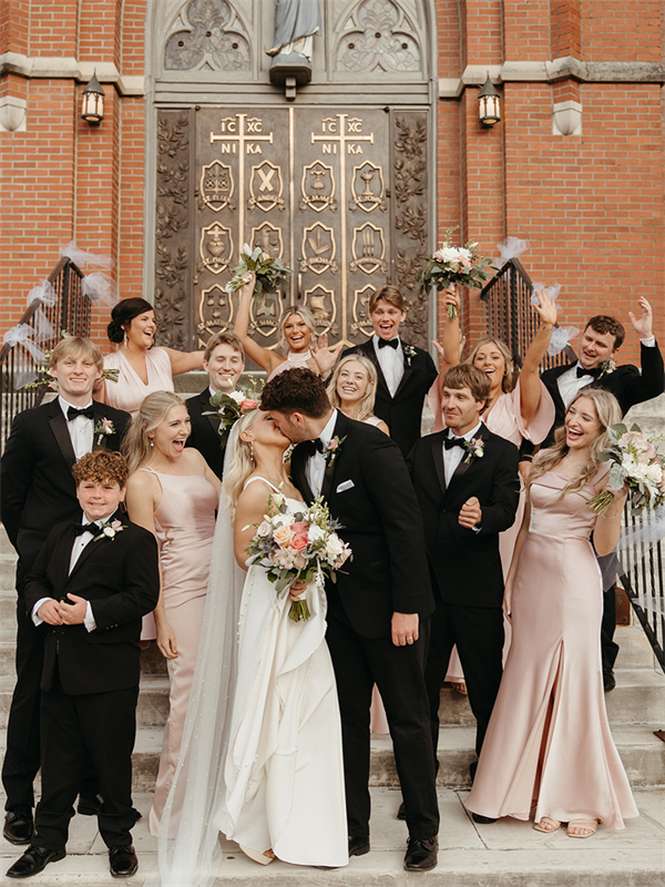 Bride and groom kissing on church steps with bridal party celebrating.