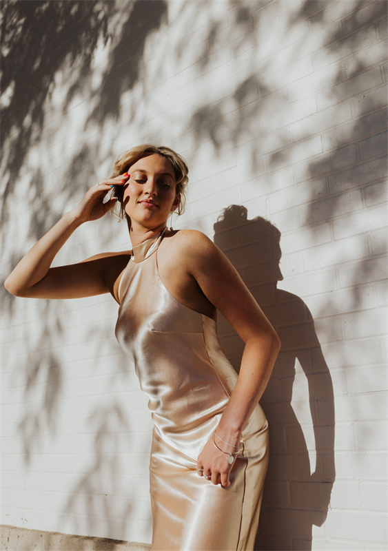 A bridesmaid in a satin dress posing against a white brick wall with leaf shadows.