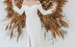 A bride in a white off-shoulder wedding gown stands in front of a decorative arrangement of dried grasses and flowers.