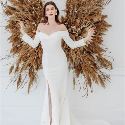 A bride in a white off-shoulder wedding gown stands in front of a decorative arrangement of dried grasses and flowers.