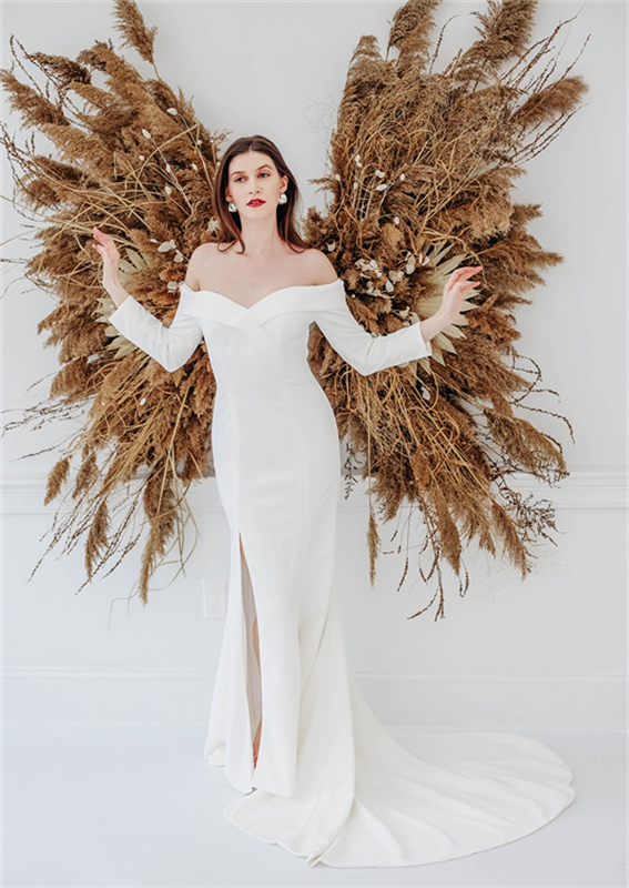 A bride in a white off-shoulder wedding gown stands in front of a decorative arrangement of dried grasses and flowers.