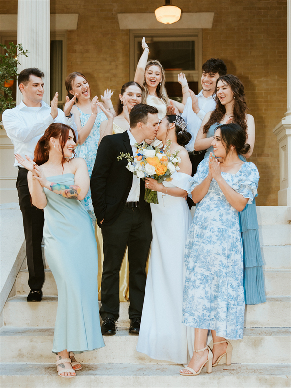 A newlywed couple kissing on the steps, surrounded by cheering and clapping friends.