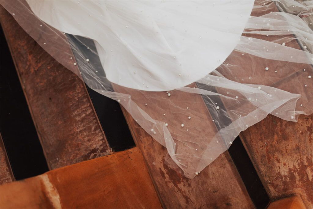 The delicate pearl-adorned fabric of the wedding dress contrasts beautifully with the rustic background.