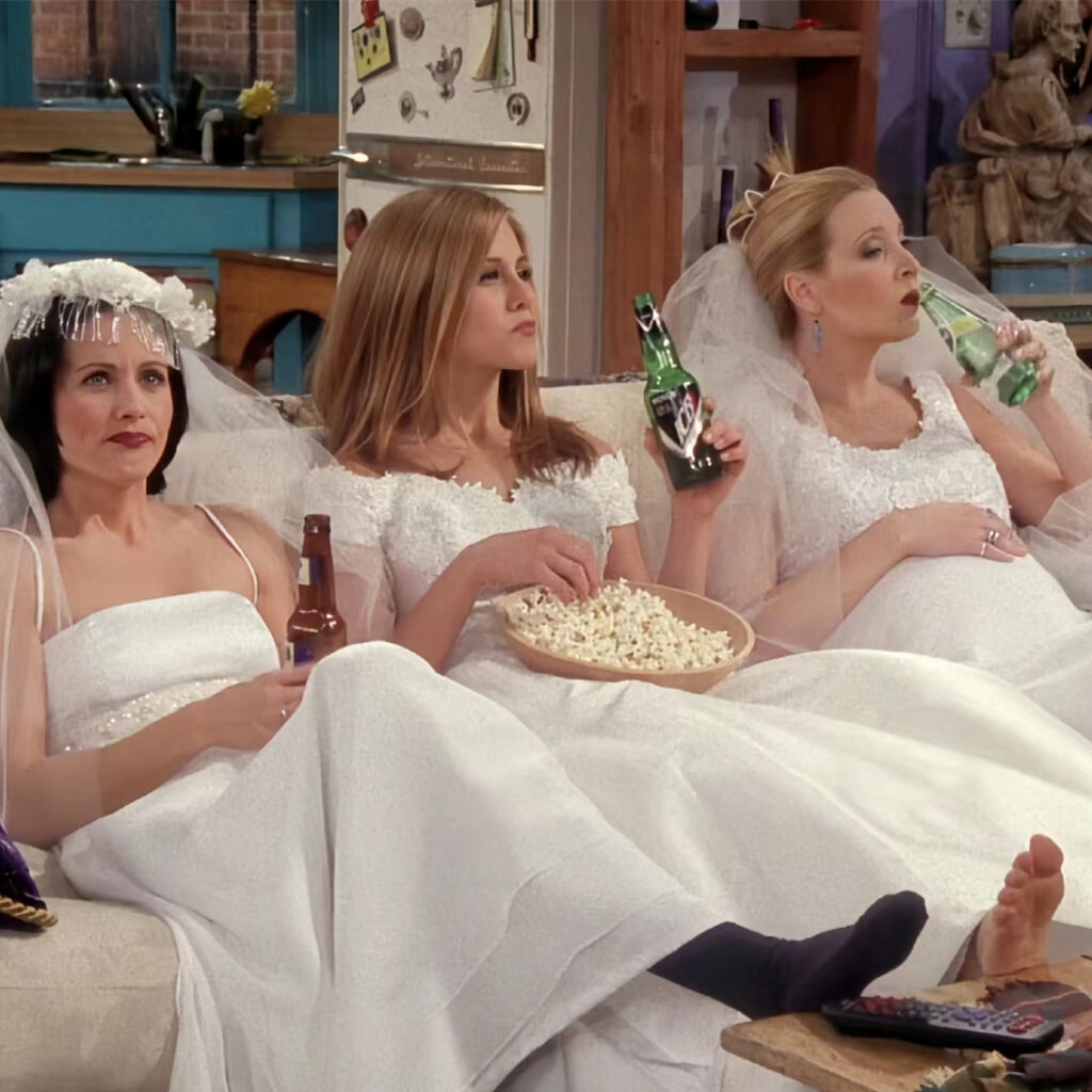 Monica, Rachel, and Phoebe from "Friends" sitting on a couch in wedding dresses.