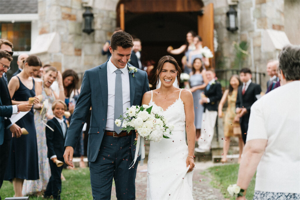 A smiling couple walk down the aisle outside, surrounded by cheering guests.