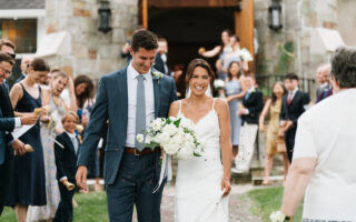 A smiling couple walk down the aisle outside, surrounded by cheering guests.