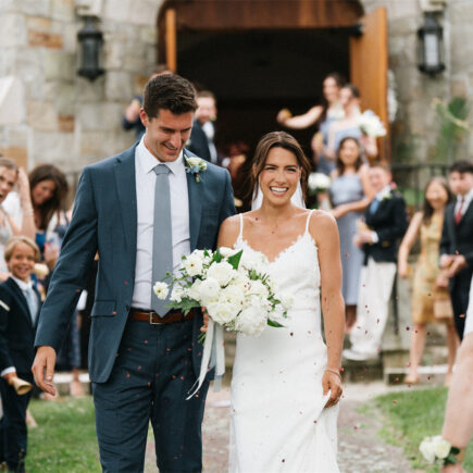 A smiling couple walk down the aisle outside, surrounded by cheering guests.