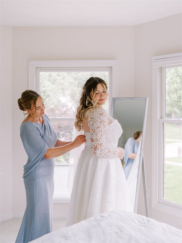 A woman helps a bride with her wedding dress in front of a mirror.