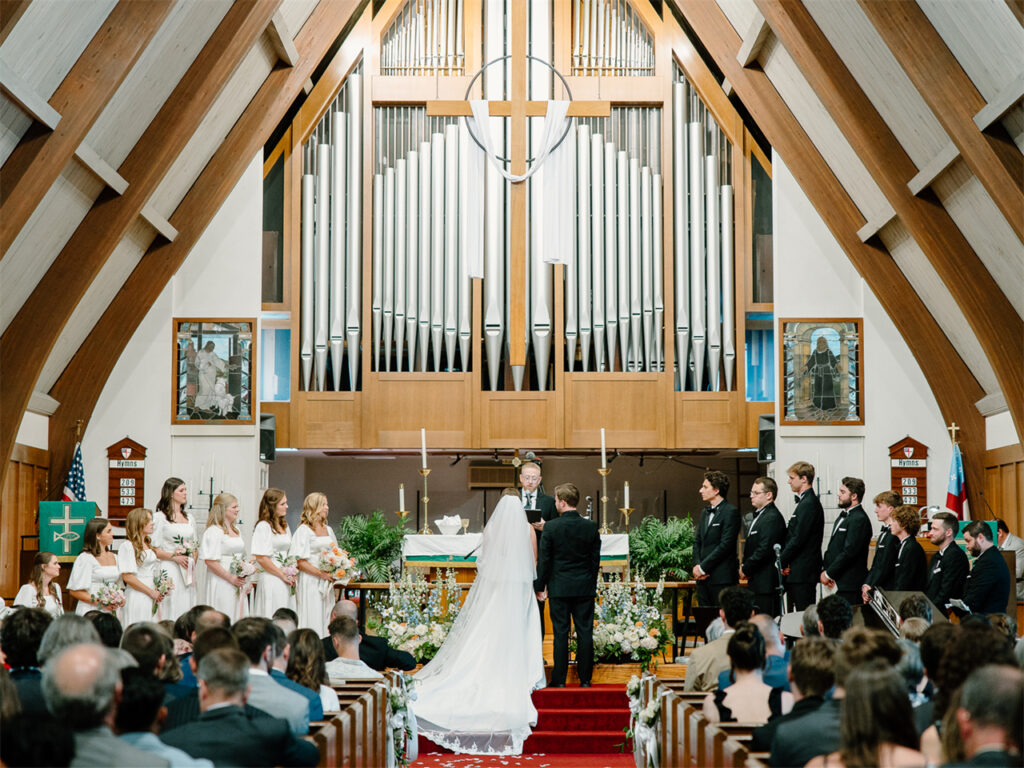 A couple stands at the altar in a church, surrounded by bridesmaids and groomsmen during their wedding ceremony.