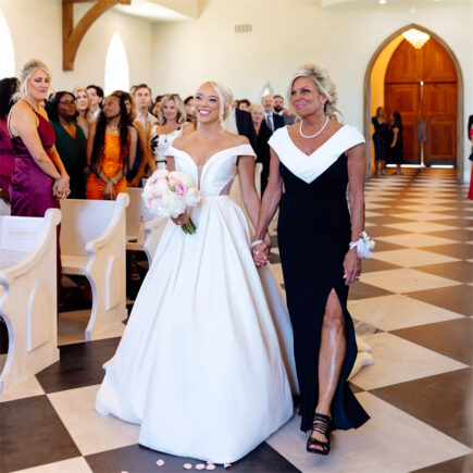 The bride and the mother of the bride walking down the aisle in a church, surrounded by guests.