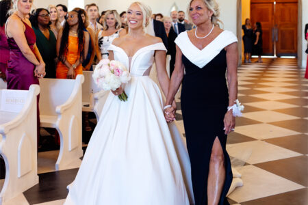 The bride and the mother of the bride walking down the aisle in a church, surrounded by guests.