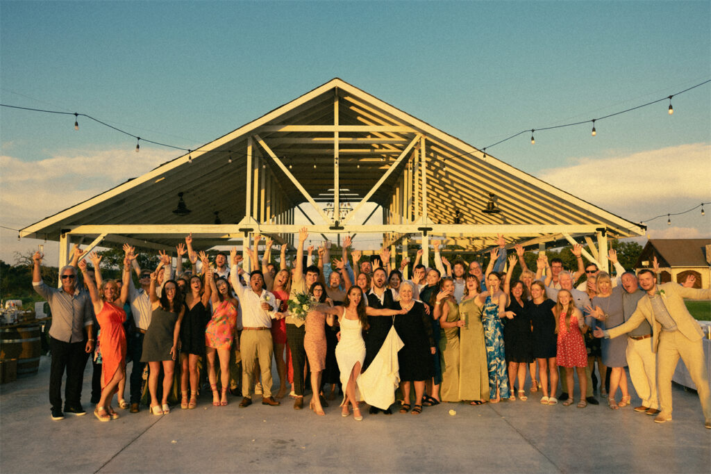 A group of people celebrating a wedding in front of a barn with hands raised.